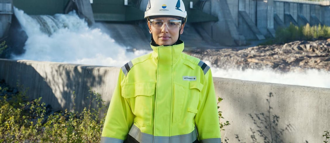 Woman in high-visibility safety clothing and helmet standing in front of a hydro power plant.