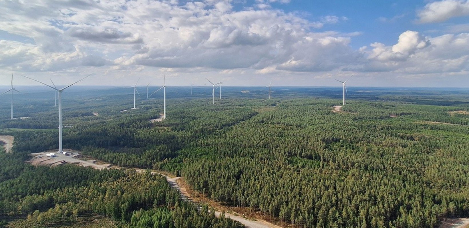 Aerial view of wind turbines spaced across dense forest, with service roads and clearings under a partly cloudy sky.
