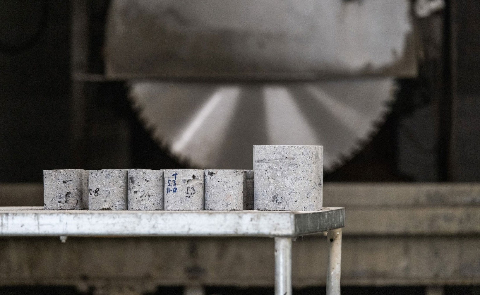 Concrete test cylinders arranged on a table, with a large rotating saw in the background, prepared for material strength testing and analysis. Concrete test cylinders arranged on a table, with a large rotating saw in the background, prepared for material strength testing and analysis.
