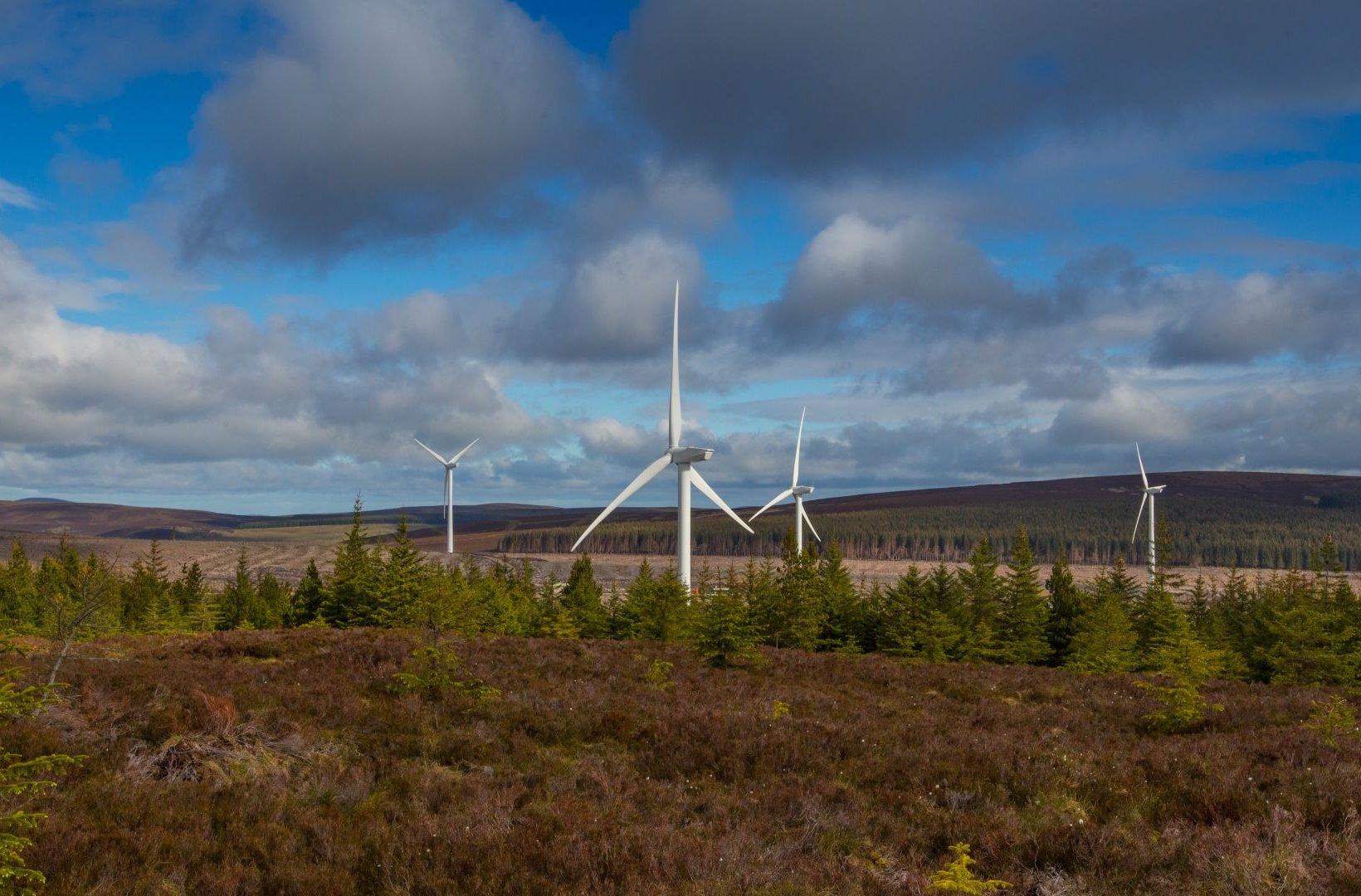 Wind turbines on a forested hillside under a partly cloudy sky.