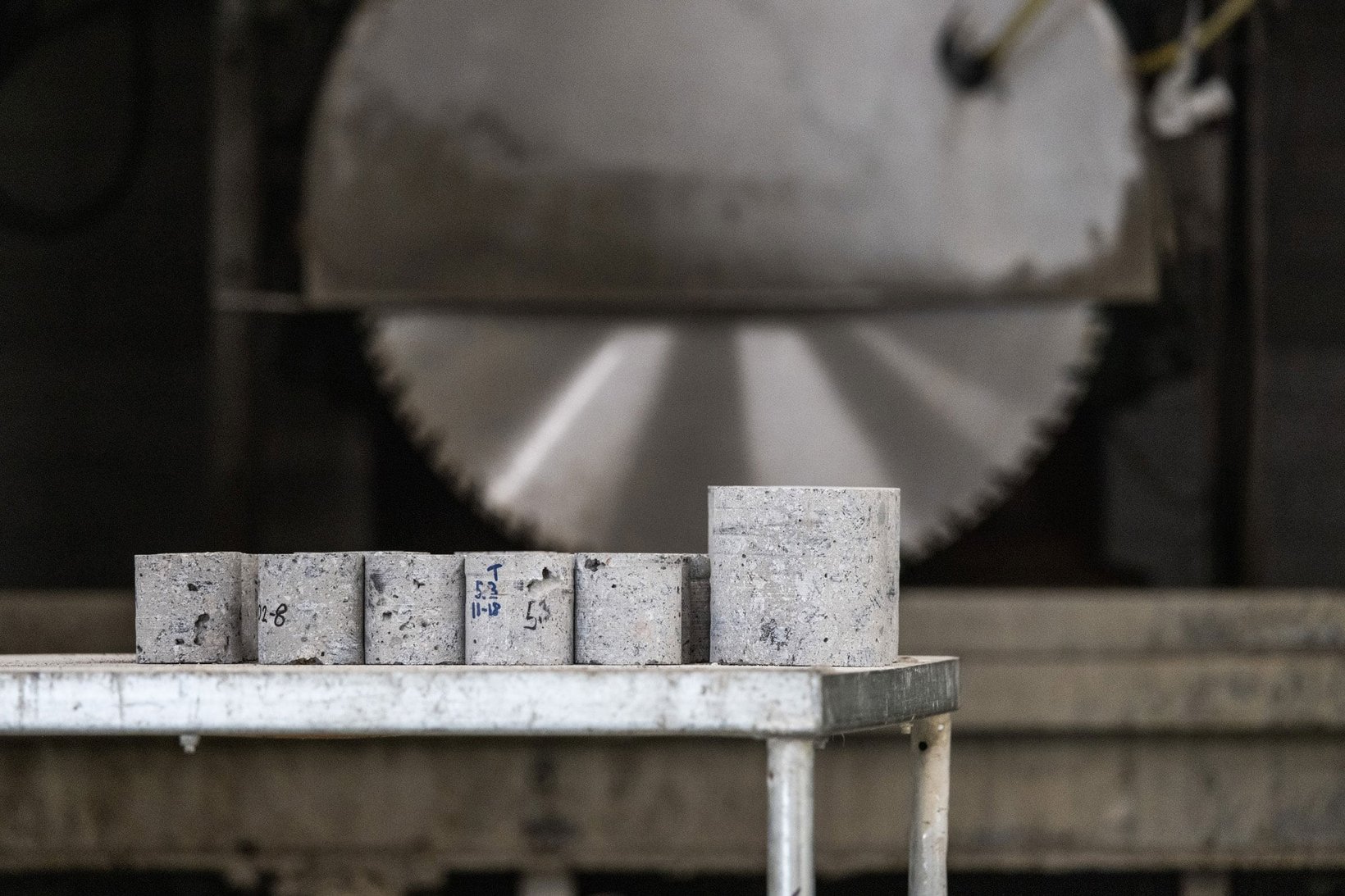 Concrete test cylinders arranged on a table, with a large rotating saw in the background, prepared for material strength testing and analysis. Concrete test cylinders arranged on a table, with a large rotating saw in the background, prepared for material strength testing and analysis.