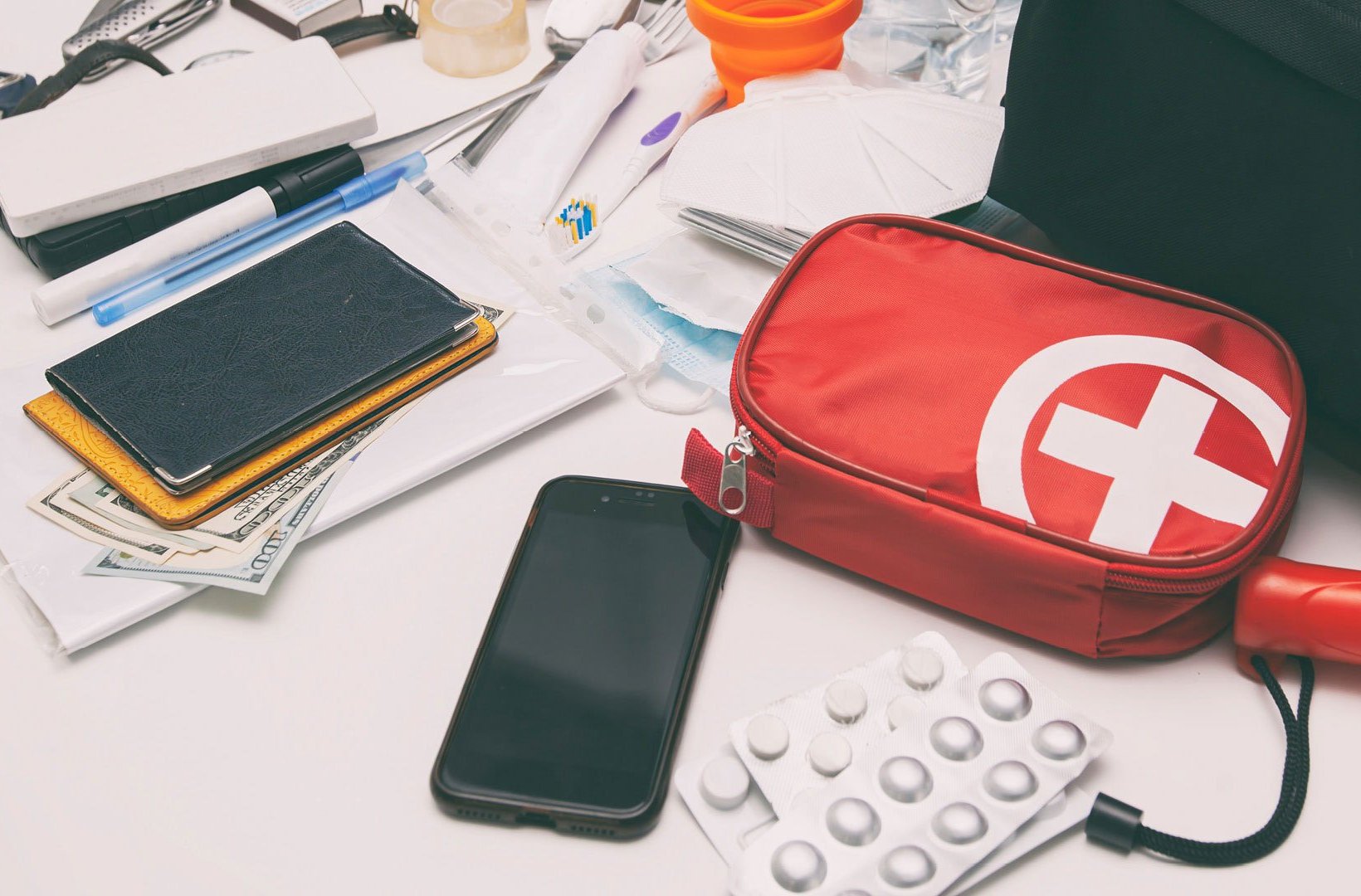 A collection of emergency and travel essentials spread out on a white surface, including a red first‑aid pouch with a white cross, blister packs of pills, a smartphone, passports resting on cash, and various small supplies such as pens, tape, and medical items.