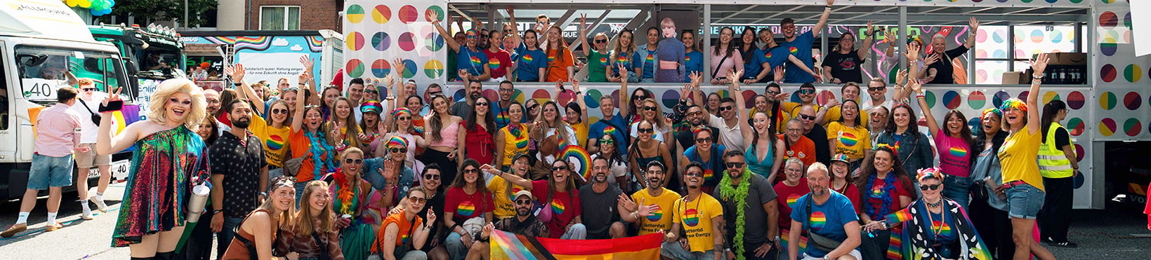 Large group gathered in front of a decorated stage, holding rainbow flags.