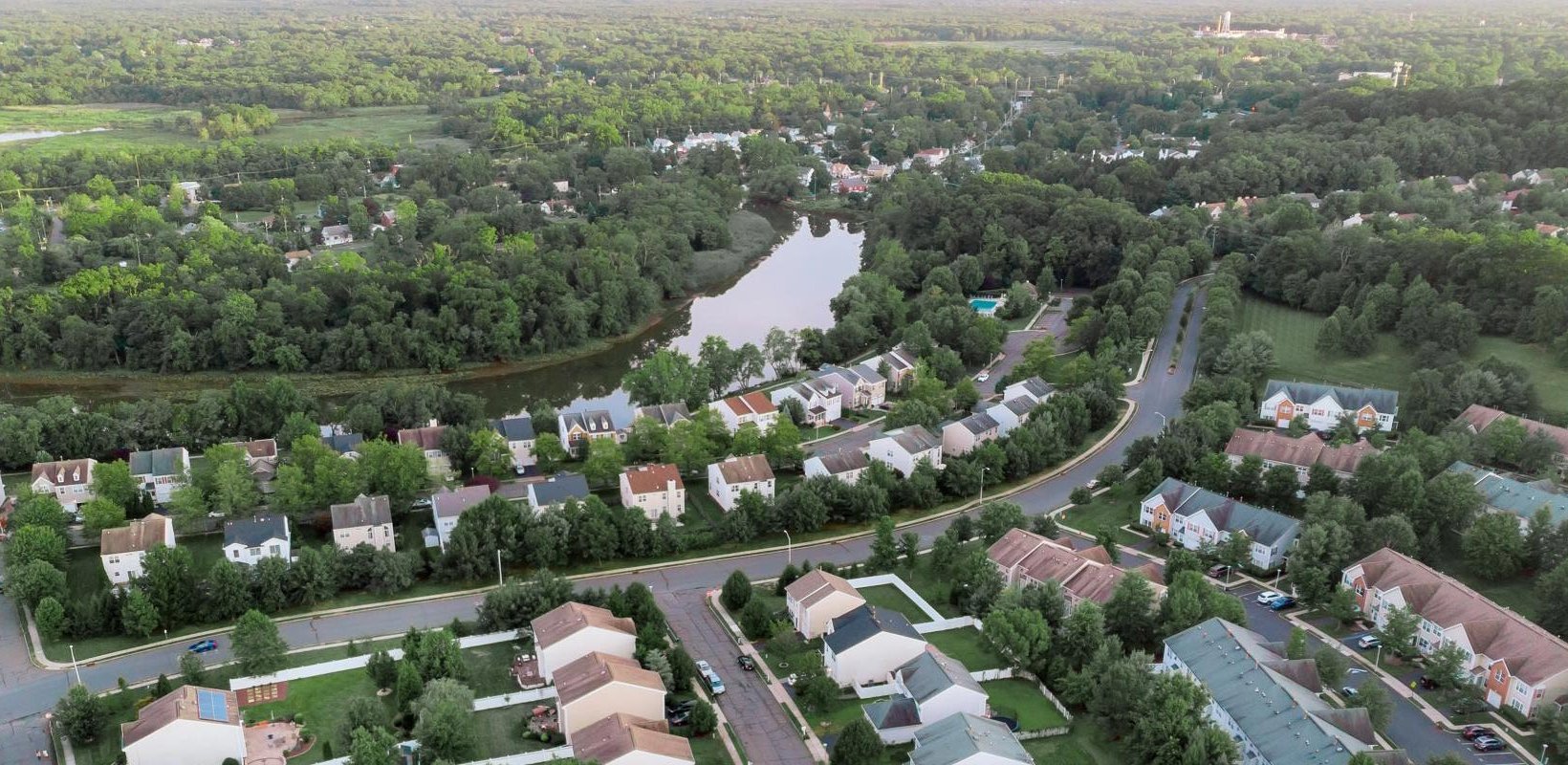 Aerial view of a leafy suburban neighbourhood with houses, winding roads, a pond, and surrounding woodland.