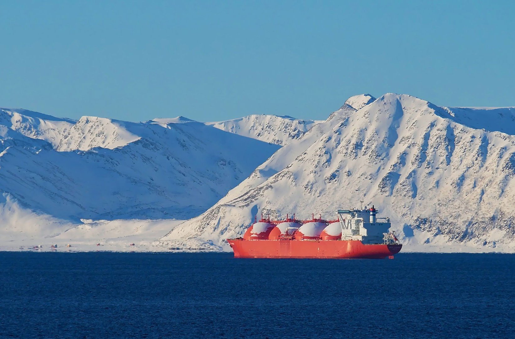 A large bright‑orange liquefied natural gas (LNG) tanker floats on deep blue water in front of a backdrop of tall, snow‑covered mountains under a clear blue sky. The rounded white storage domes on the ship stand out against the icy landscape. A large bright‑orange liquefied natural gas (LNG) tanker floats on deep blue water in front of a backdrop of tall, snow‑covered mountains under a clear blue sky. The rounded white storage domes on the ship stand out against the icy landscape.