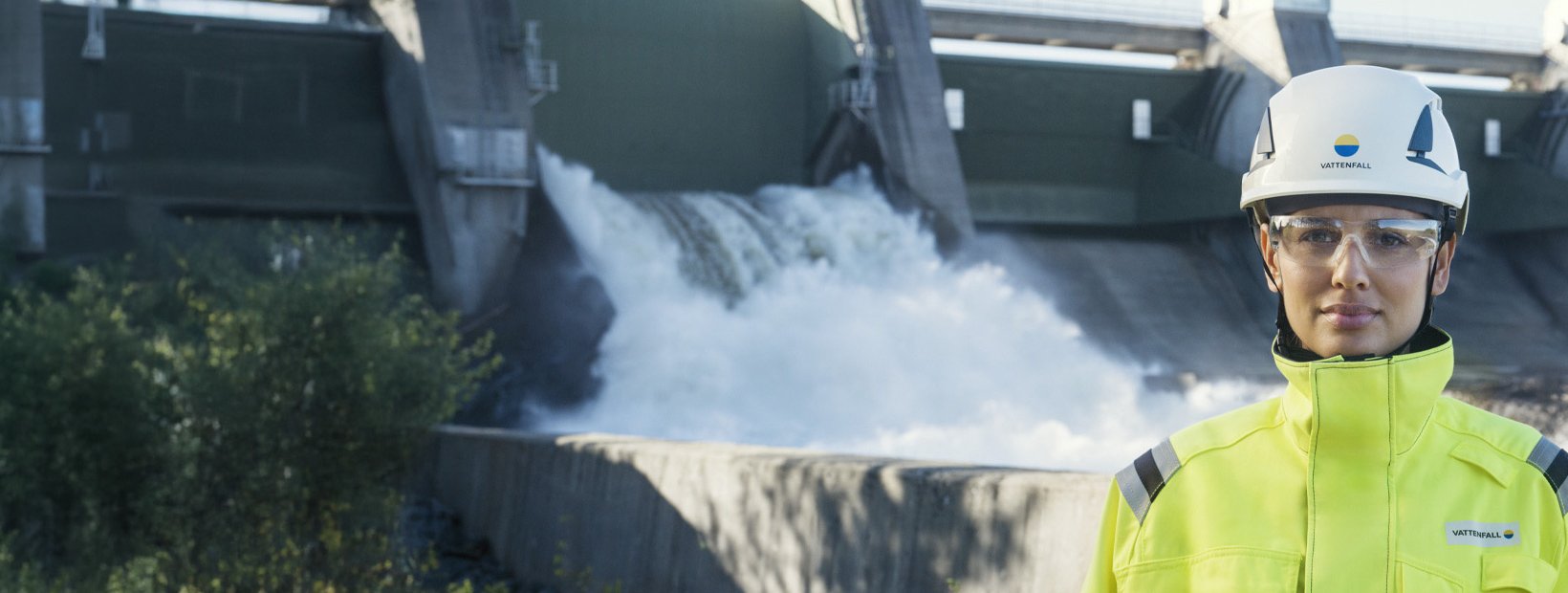 Woman in high-visibility safety clothing and helmet standing in front of a hydro power plant.