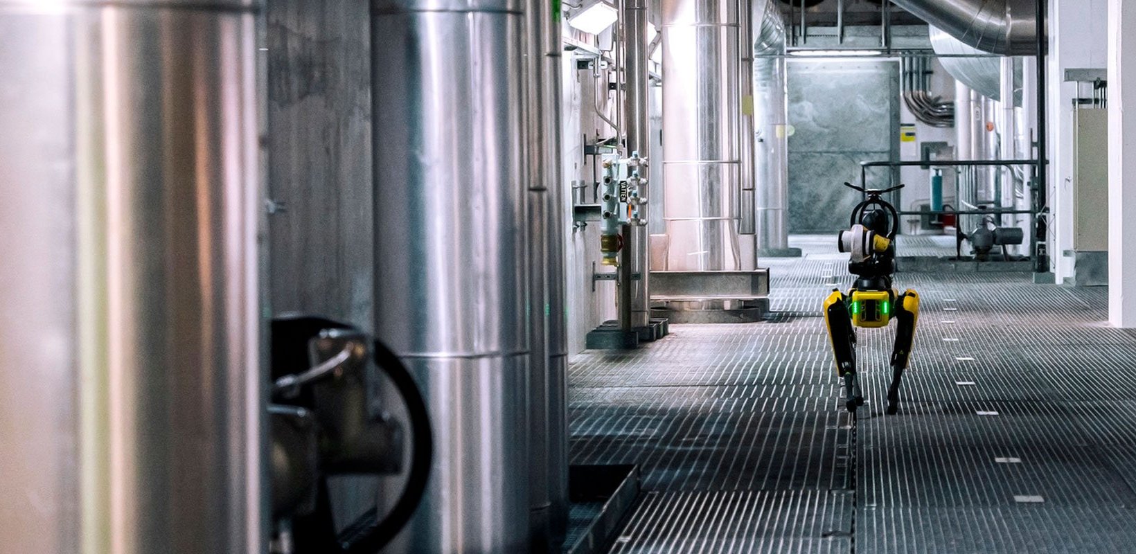 The robot dog Spot walking along a metal-grated floor inside an industrial facility with large pipes, ducts, and stainless-steel equipment.