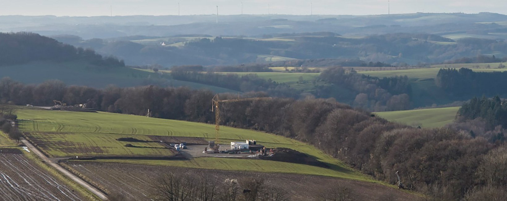 Aerial view of a rural construction site with a tall yellow tower crane, surrounded by fields and woodland. Aerial view of a rural construction site with a tall yellow tower crane, surrounded by fields and woodland.