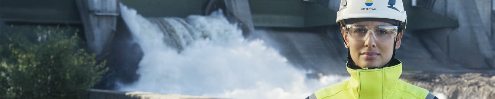 Woman in high-visibility safety clothing and helmet standing in front of a hydro power plant.