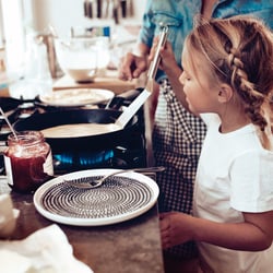 Child standing beside a kitchen hob while pancakes cook in a frying pan over a blue gas flame.