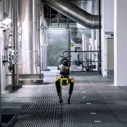 The robot dog Spot walking along a metal-grated floor inside an industrial facility with large pipes, ducts, and stainless-steel equipment.