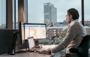 Man working at a desk with multiple screens beside a large window.