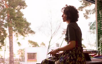 A woman sitting indoors by a large window, looking out toward trees and houses. A woman sitting indoors by a large window, looking out toward trees and houses.