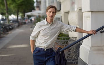 Young man wearing a light shirt and blue trousers leaning on a stair railing beside a city street.