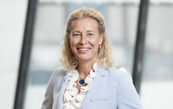 Annika Ramsköld, Head of Sustainability at Vattenfall, wearing a light blue blazer and a white blouse, standing indoors in front of large windows in a modern office environment.