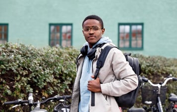 A young man with a backpack standing in front of parked bicycles and a green building.