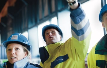 Workers in helmets and high‑visibility jackets look up; one points inside a building.