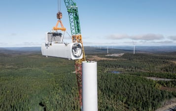 Crane lifting a wind turbine nacelle into position on top of a tower, with forested landscape and other turbines in the background.
