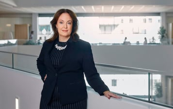 Anna Borg, Vattenfall's President and CEO, stands by a railing in a modern office interior, wearing a dark blazer and a striped dress.