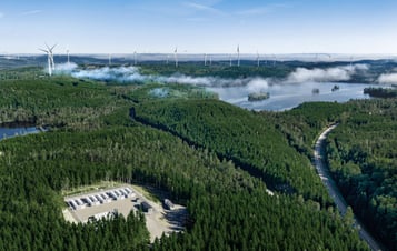 A forested landscape with wind turbines, a battery array, lakes and a road.