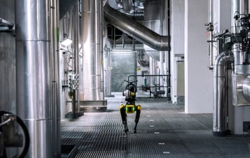 The robot dog Spot walking along a metal-grated floor inside an industrial facility with large pipes, ducts, and stainless-steel equipment.