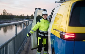 A woman in high‑visibility clothing next to a service van beside a body of water. A woman in high‑visibility clothing next to a service van beside a body of water.