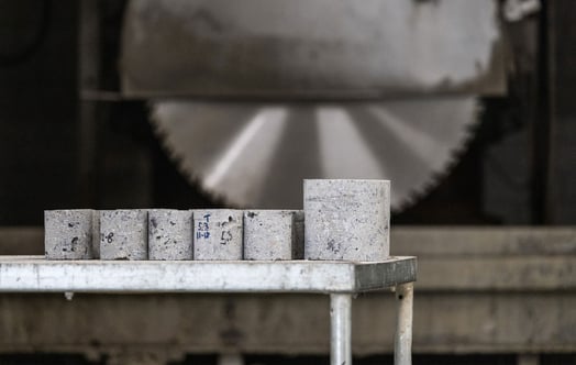 Concrete test cylinders arranged on a table, with a large rotating saw in the background, prepared for material strength testing and analysis. Concrete test cylinders arranged on a table, with a large rotating saw in the background, prepared for material strength testing and analysis.