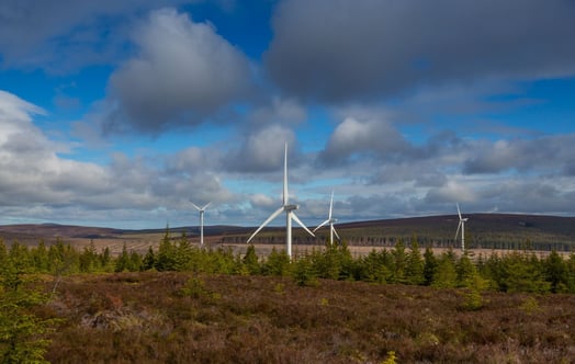 Wind turbines on a forested hillside under a partly cloudy sky.
