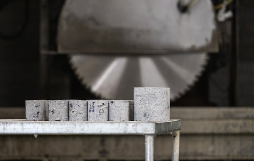 Concrete test cylinders arranged on a table, with a large rotating saw in the background, prepared for material strength testing and analysis.