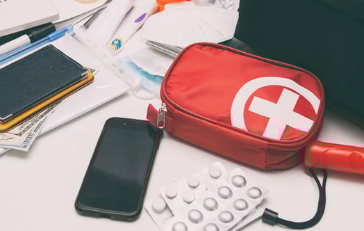 A collection of emergency and travel essentials spread out on a white surface, including a red first‑aid pouch with a white cross, blister packs of pills, a smartphone, passports resting on cash, and various small supplies such as pens, tape, and medical items.