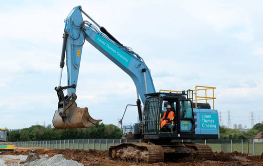 A “Lower Thames Crossing” branded excavator at a construction site, with a worker in high-visibility clothing seated in the cab. A “Lower Thames Crossing” branded excavator at a construction site, with a worker in high-visibility clothing seated in the cab.