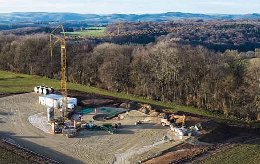 Aerial view of a rural construction site with a tall yellow tower crane, construction vehicles and temporary buildings, surrounded by fields, woodland, and rolling hills in the background. Aerial view of a rural construction site with a tall yellow tower crane, construction vehicles and temporary buildings, surrounded by fields, woodland, and rolling hills in the background.