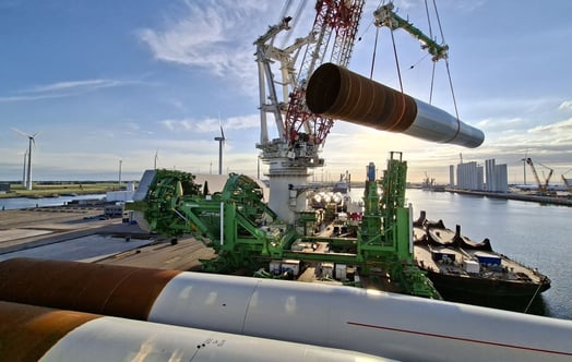 A large crane lifting a monopile foundation at a port, with installation equipment and wind turbines visible nearby.