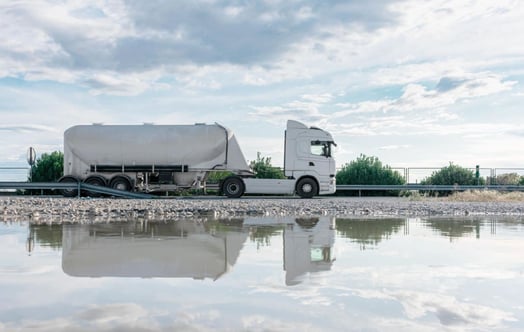 A white tanker truck driving along a road, reflected in the stream beside it.