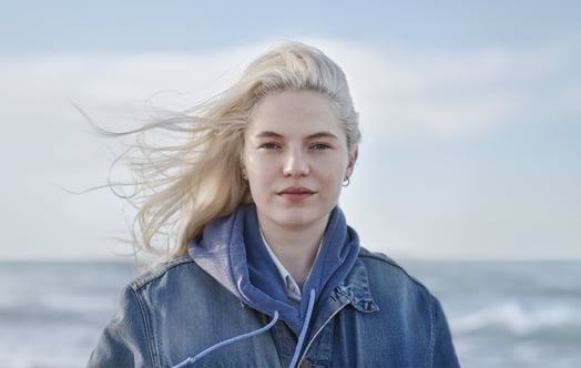 Woman in a blue jacket by the sea, with wind‑blown light hair.