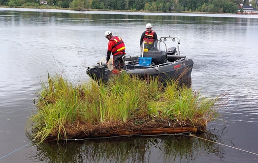 Floating island with vegetation in a river, positioned in front of a small work boat. Photo: Maidul Choudhury.