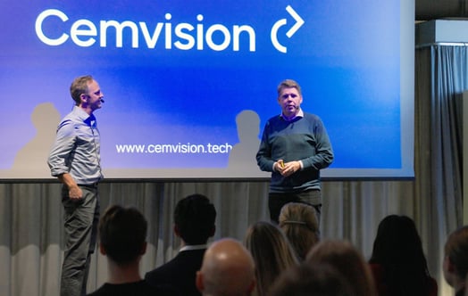 Two people stand on a stage in front of a presentation screen displaying “Cemvision”, with an audience seated in the foreground. Two people stand on a stage in front of a presentation screen displaying “Cemvision”, with an audience seated in the foreground.