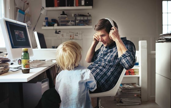 Man at a desk in a home office taking off his headphones while a child stands nearby.