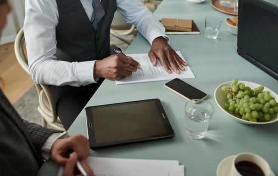 Person signing documents at a table with digital devices nearby.