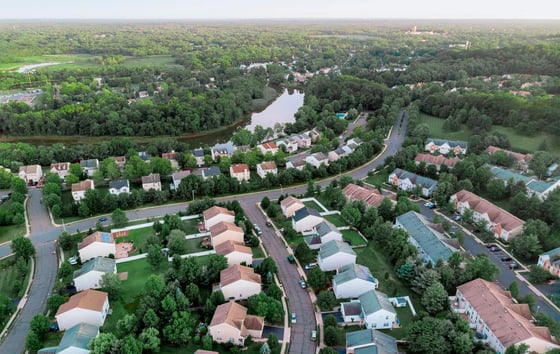 Aerial view of a leafy suburban neighbourhood with houses, winding roads, a pond, and surrounding woodland.