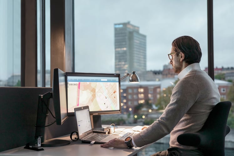 Man working at a desk with multiple screens beside a large window. Man working at a desk with multiple screens beside a large window.