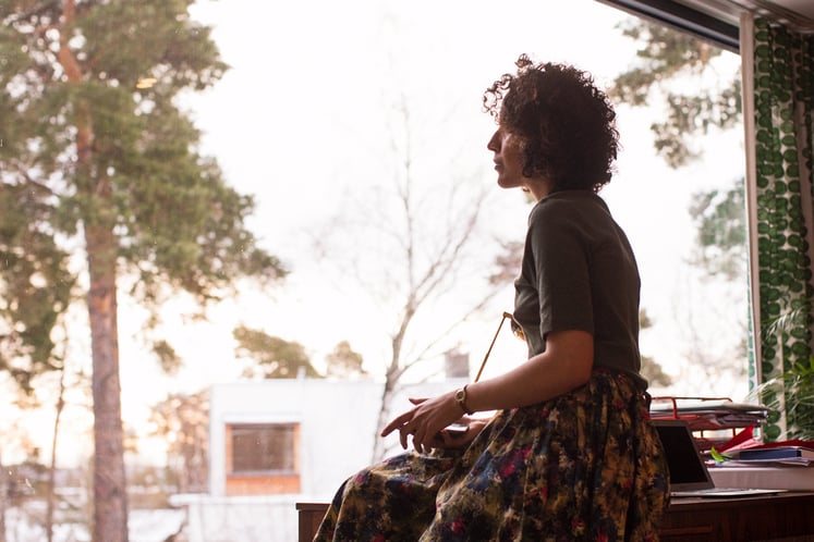A woman sitting indoors by a large window, looking out toward trees and houses.