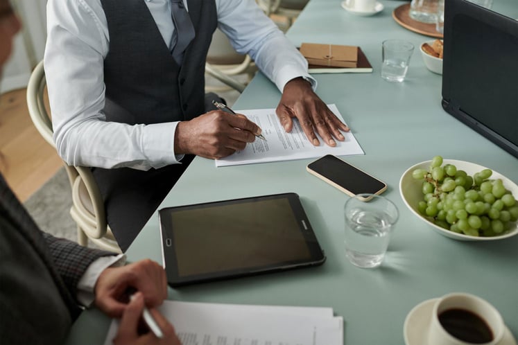 Person signing documents at a table with digital devices nearby. Person signing documents at a table with digital devices nearby.