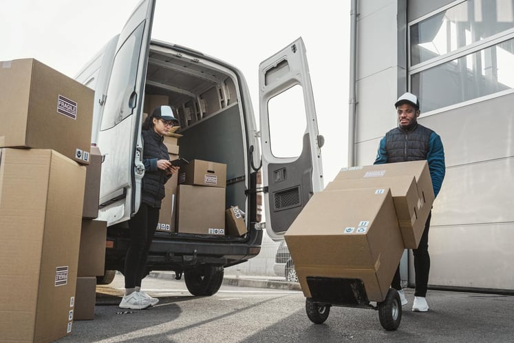 Two workers unloading cardboard boxes from a van outside a warehouse.