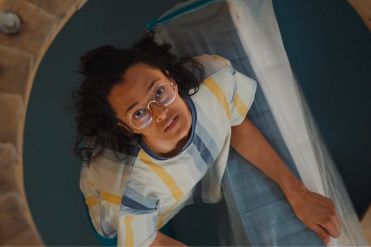 A worried young woman carrying a mattress looks up a tall spiral staircase.
