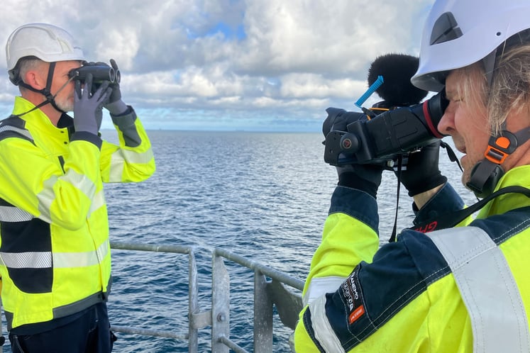 Two people wearing high-visibility clothing and safety helmets standing on a vessel at sea, using binoculars and camera equipment to observe and document offshore conditions.