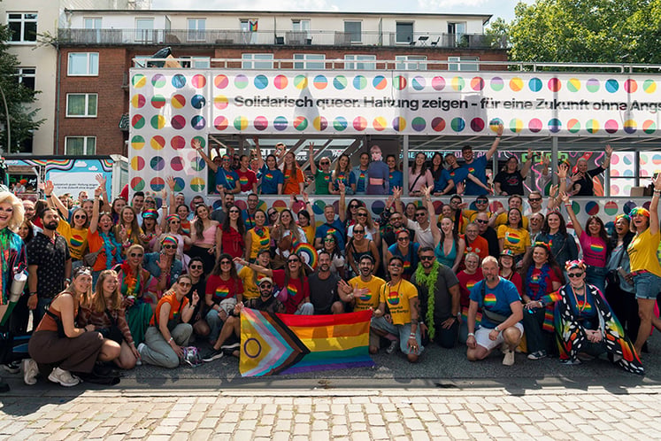 Large group gathered in front of a decorated stage, holding rainbow flags.