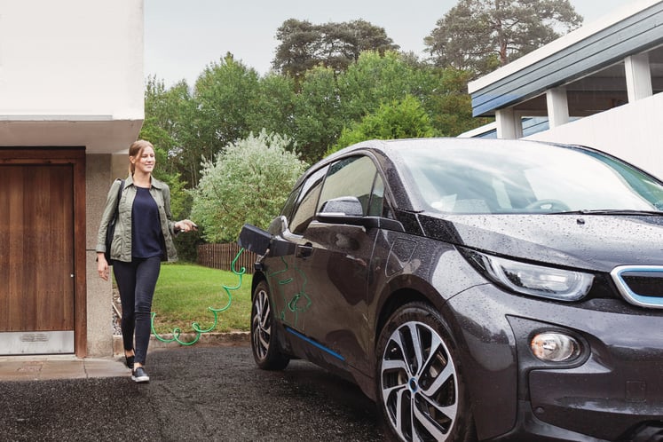 A woman walking towards an electric car being charged outside a house.