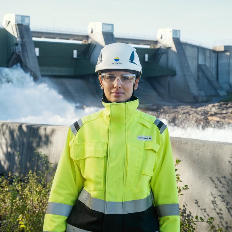 Woman in high-visibility safety clothing and helmet standing in front of a hydro power plant.