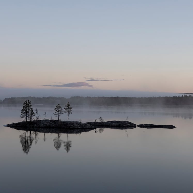 Small rocky islets with sparse pine trees in a calm lake, light mist and reflections under a pale sky. Small rocky islets with sparse pine trees in a calm lake, light mist and reflections under a pale sky.